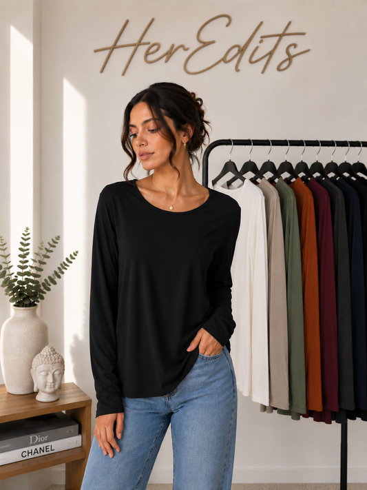 A woman wearing a black long-sleeve crewneck top and light blue jeans stands in a boutique setting with a clothing rack of other colors behind her and 'HerEdits' written on the wall.