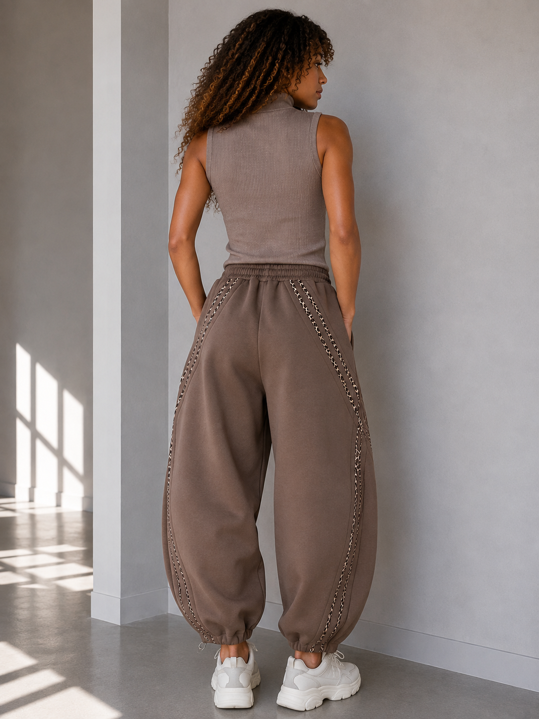 A woman shown from behind wearing loose, wide-leg taupe-brown harem-style pants with decorative chain-like trim along the sides, paired with a sleeveless top and white sneakers in a minimalist indoor setting.