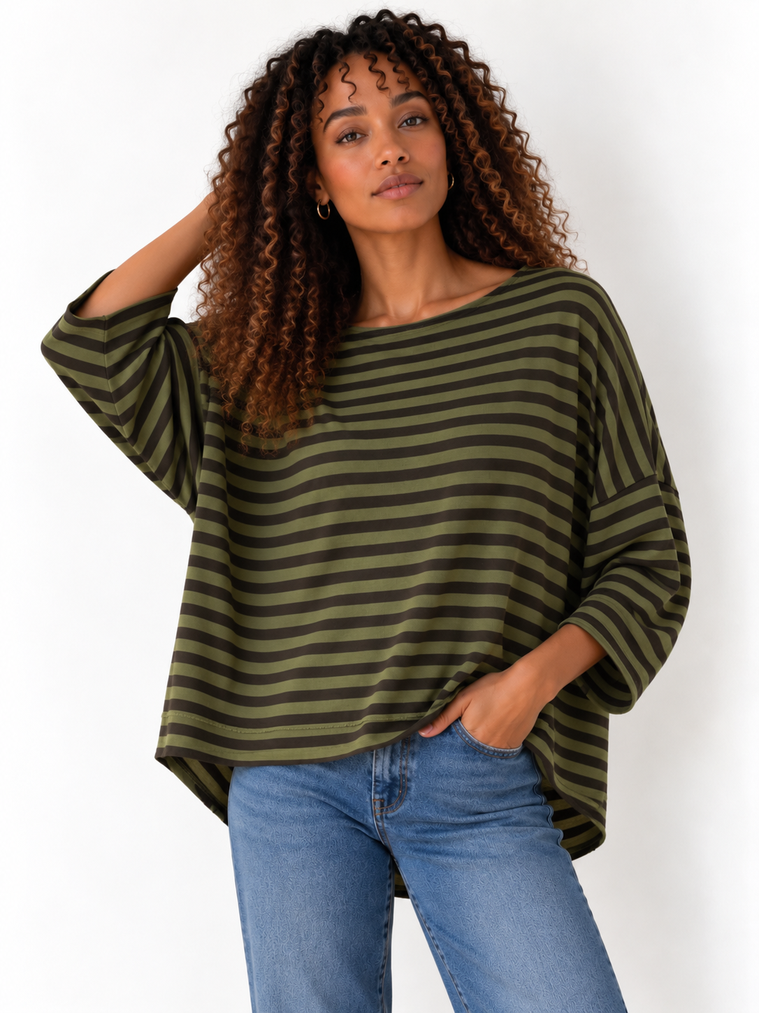 A woman with curly brown hair wearing a loose, olive green and dark green striped top with blue jeans, posing against a plain white background.