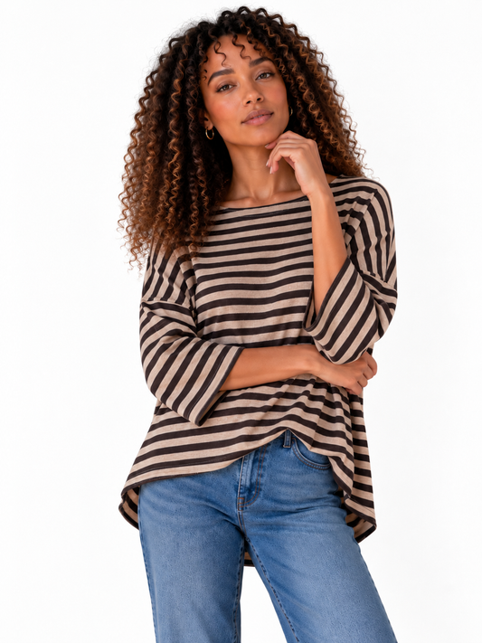 A woman with curly brown hair wearing a beige and dark brown horizontally striped boatneck top and blue jeans, standing against a white wall.