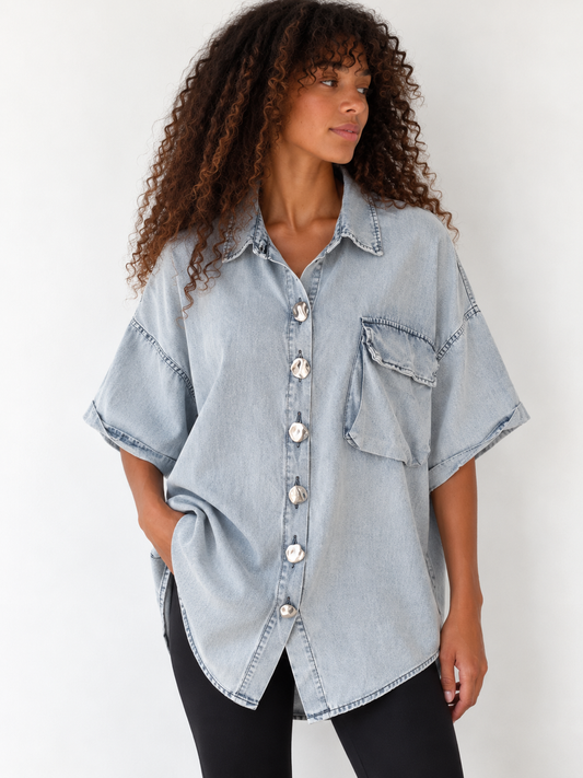 A woman wearing an oversized light-wash denim shirt with short sleeves, a button front, and a chest pocket, paired with black pants, against a plain white background.