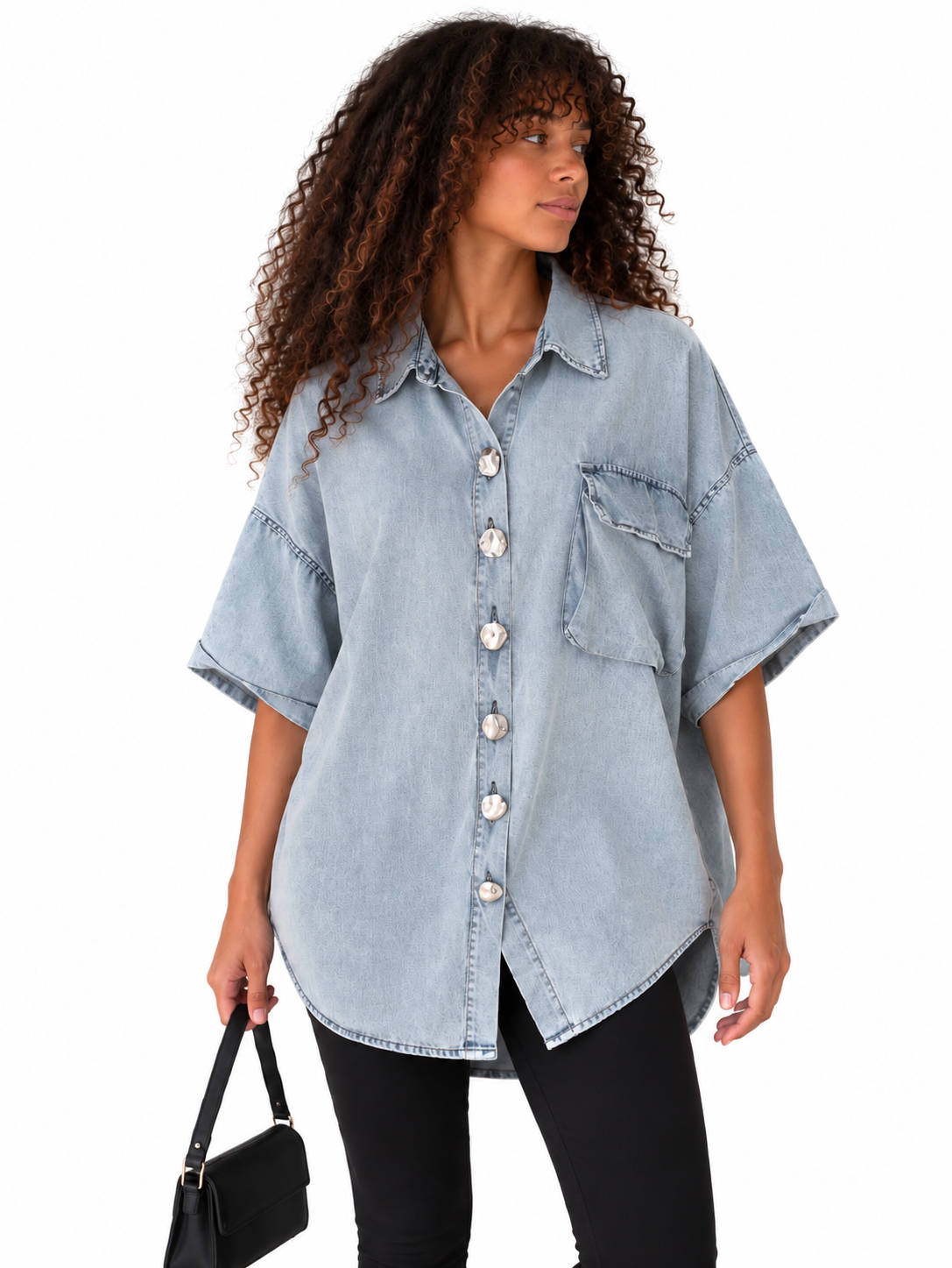 A woman modeling an oversized light blue denim button-up shirt with short sleeves, a chest pocket, and decorative stitching, paired with black pants and holding a black handbag against a plain white background.