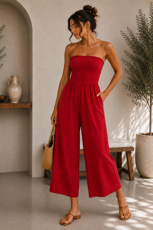 A woman models a bright red strapless wide-leg jumpsuit in a styled indoor setting with neutral walls, a wooden bench, and potted plants.
