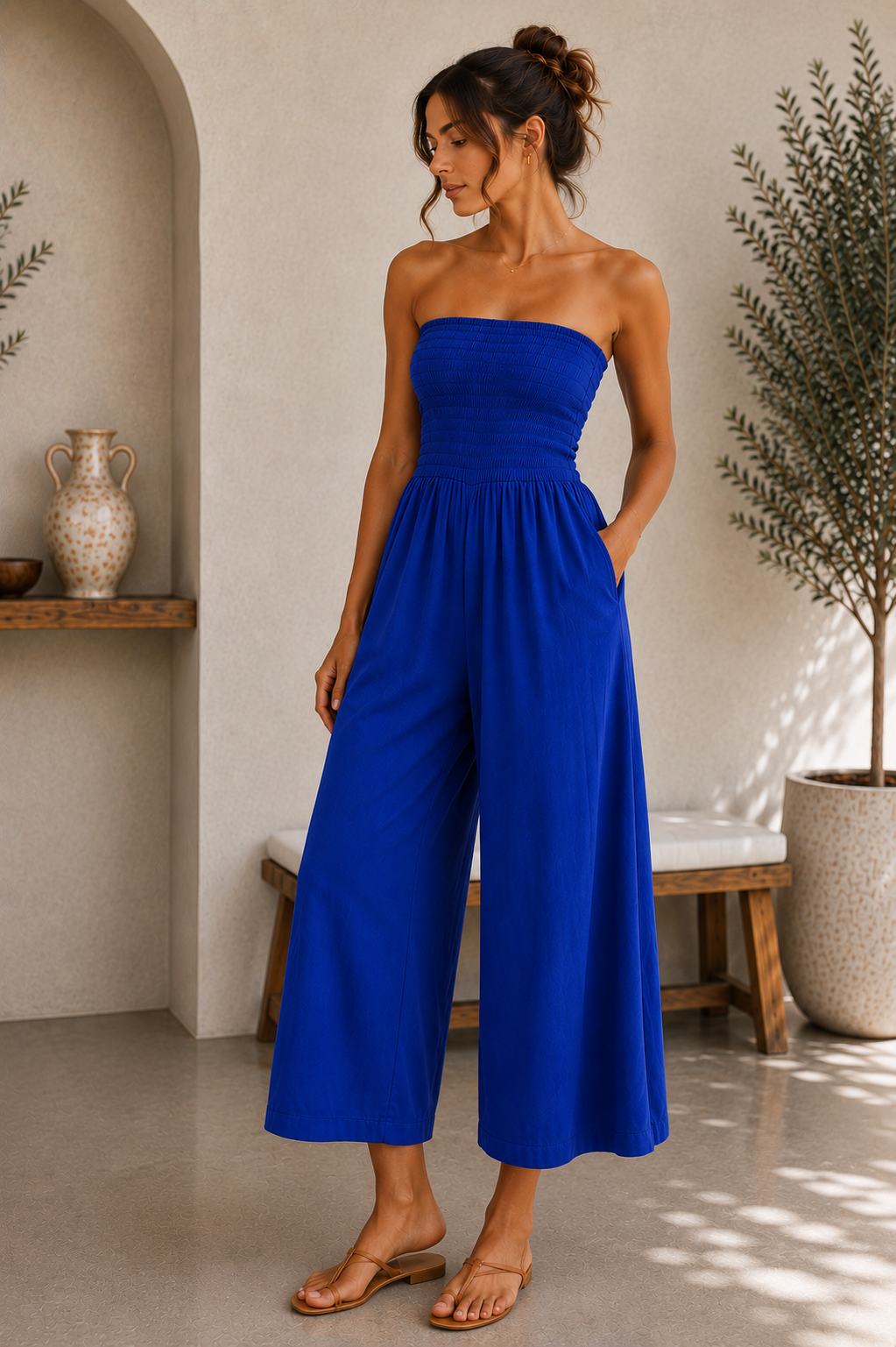A woman wearing a bright royal blue strapless wide-leg jumpsuit, standing in a sunlit indoor room with neutral walls, a bench, and potted plants.