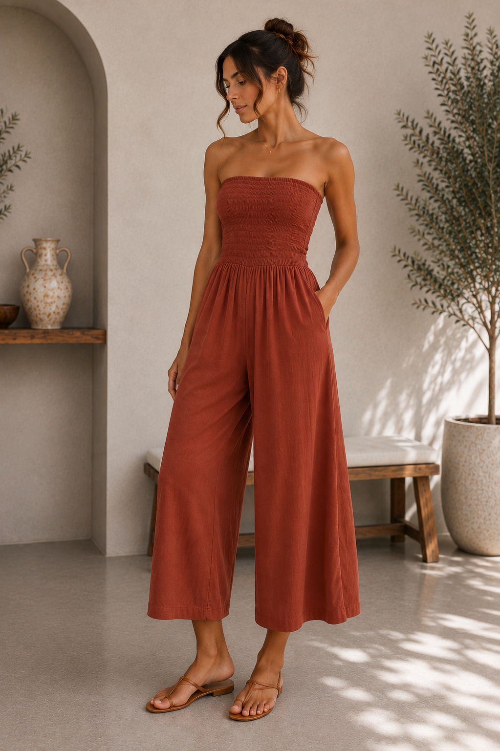 A woman models a terracotta-colored strapless wide-leg jumpsuit in a sunlit, rustic interior setting with a bench, potted plant, and textured walls.