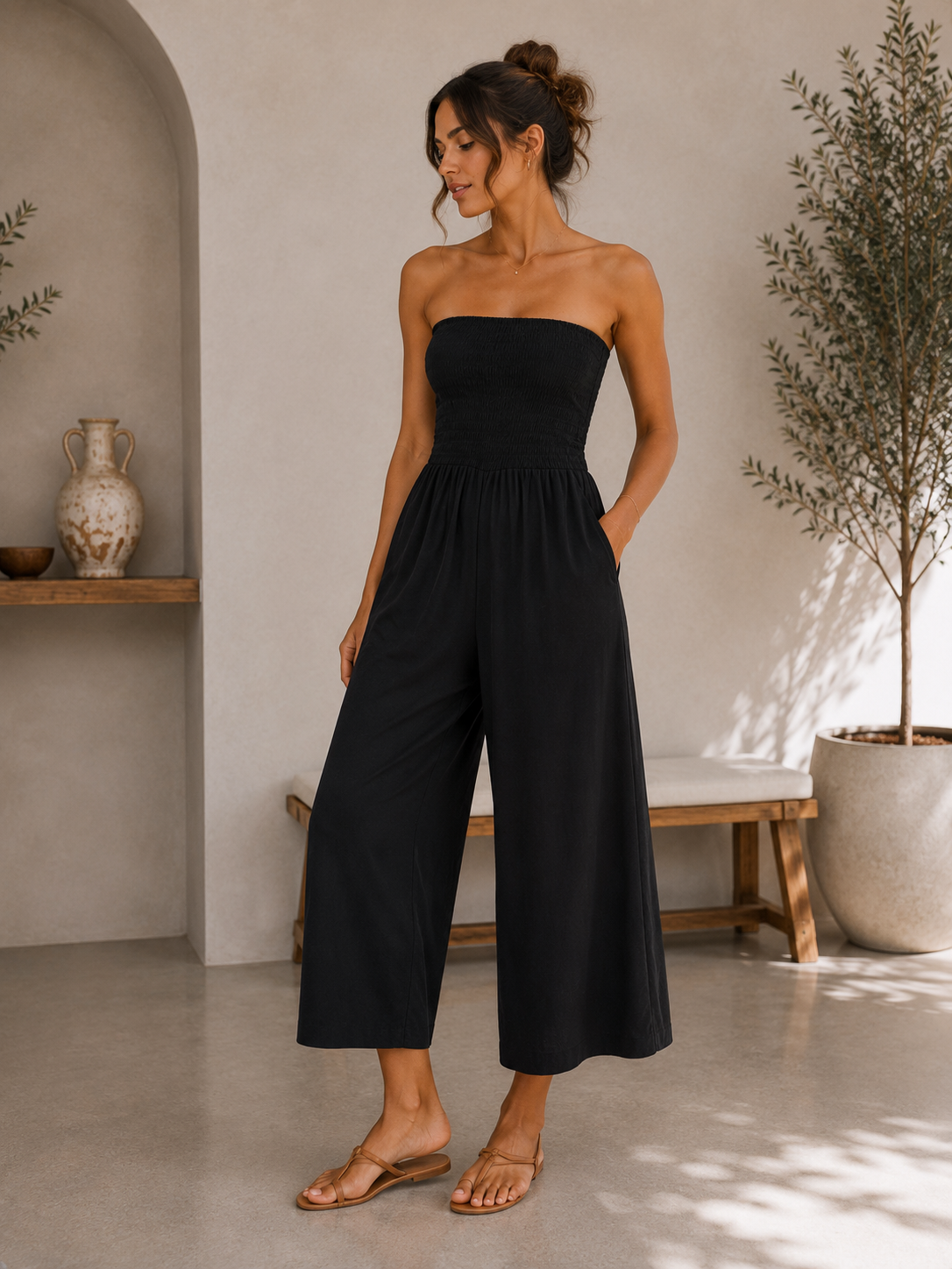 A woman modeling a strapless black wide-leg jumpsuit in a bright, modern interior setting with a bench, potted plant, and neutral-toned walls.