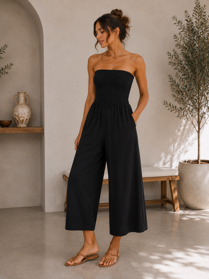 A woman modeling a strapless black wide-leg jumpsuit in a bright, modern interior setting with a bench, potted plant, and neutral-toned walls.