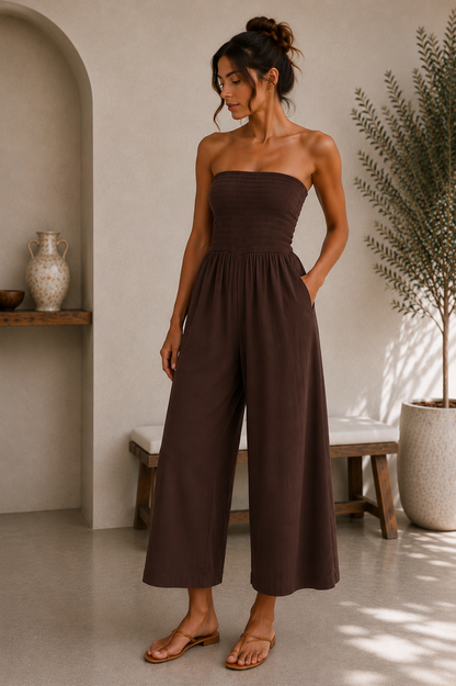 A woman wearing a dark brown strapless wide-leg jumpsuit, standing in a minimalist interior setting with a bench, potted plant, and arched alcove.