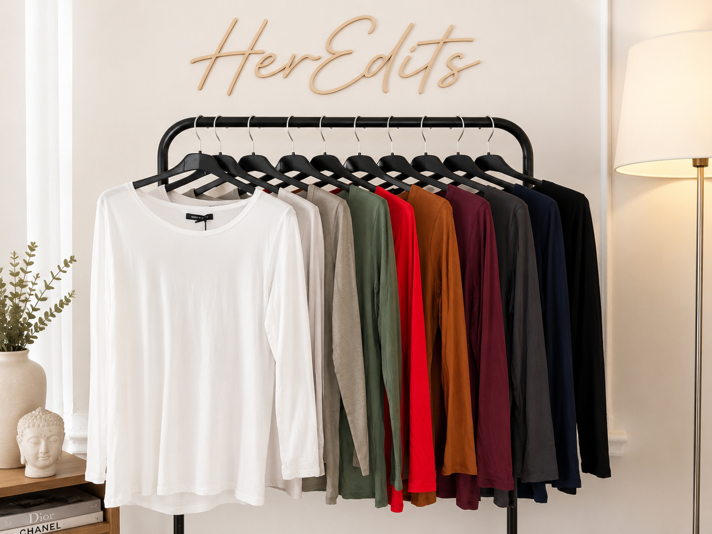 A clothing rack displaying a lineup of women's long-sleeve topshirts in various colors, including white, beige, gray, red, rust, burgundy, navy, and black. The rack is set against a warm beige wall with a wooden 'HerEdits' sign above it, flanked by a lit floor lamp and a small decorative table on the left.