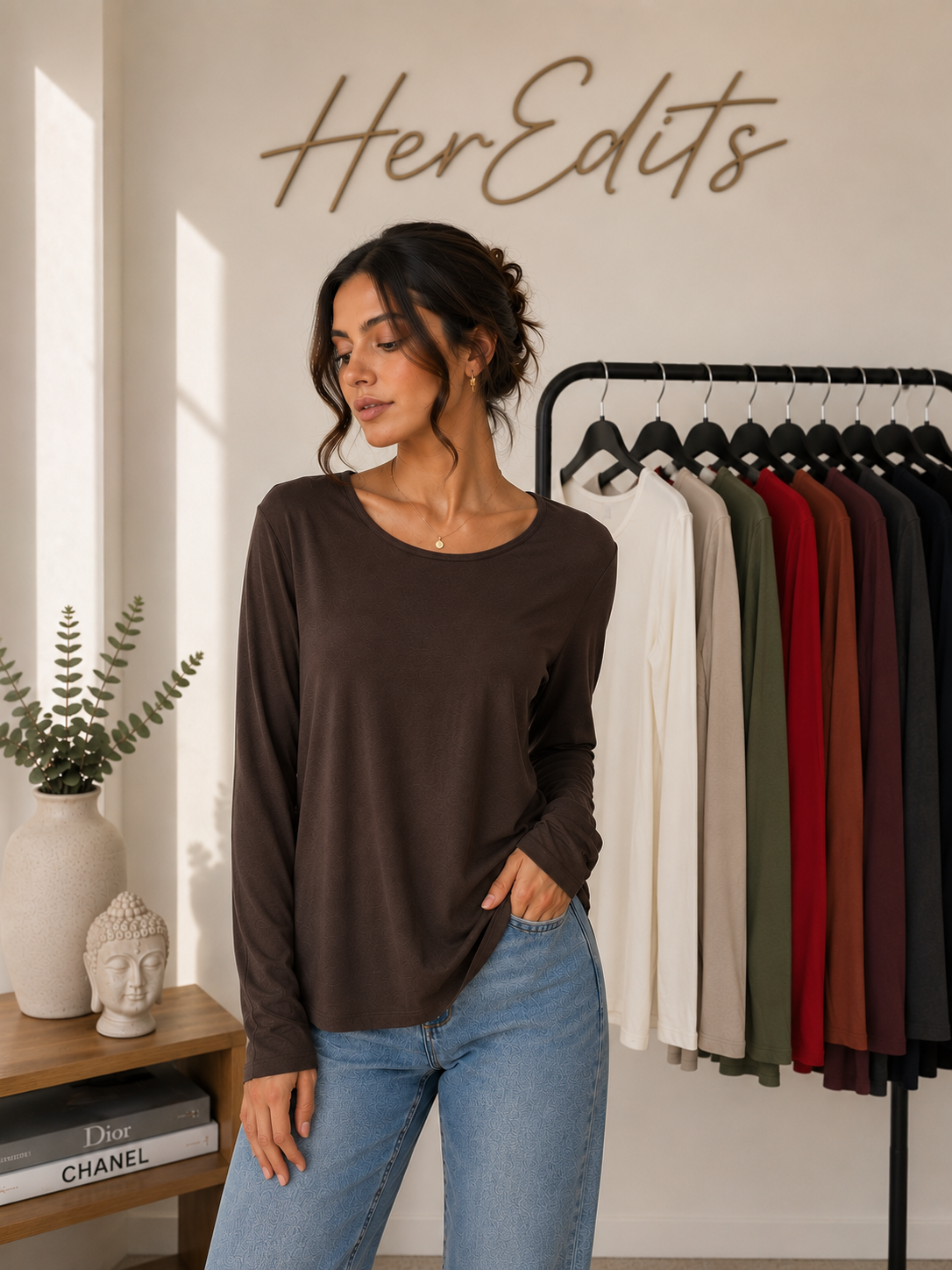 A woman wearing a dark brown long-sleeve top and light blue jeans stands in a boutique setting with a clothing rack of other colors behind her and a wall sign reading 'HerEdits'.