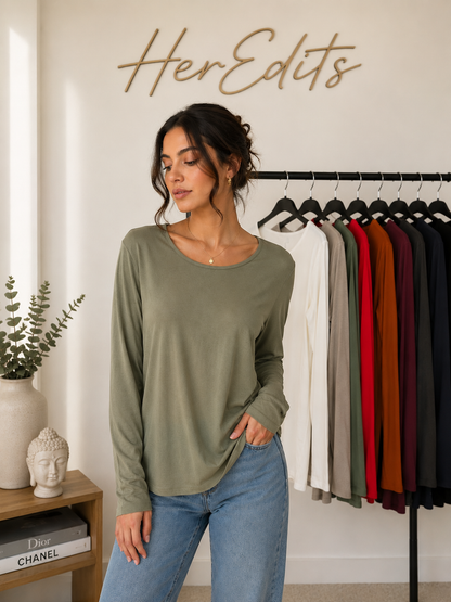 A woman wearing a sage green long-sleeve top and blue jeans stands in a boutique setting with a clothing rack of other colors behind her and a wall sign reading 'HerEdits'.