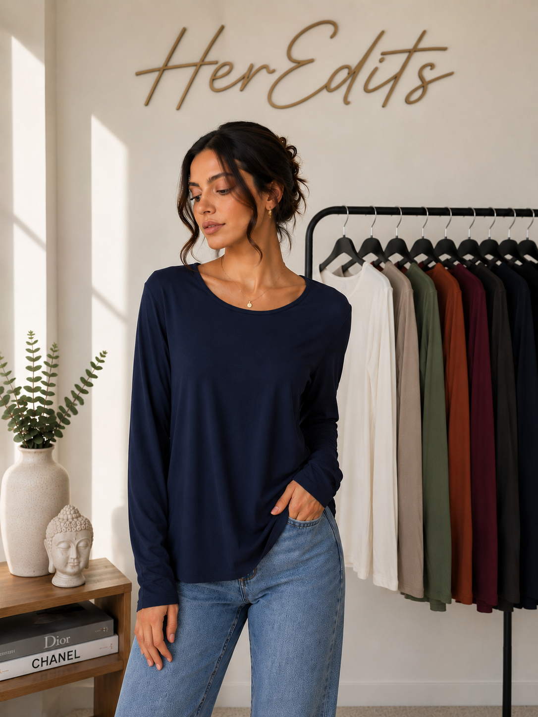 A woman wearing a navy blue long-sleeve top and light blue jeans stands in a boutique setting with a clothing rack and the words 'HerEdits' on the wall behind her.