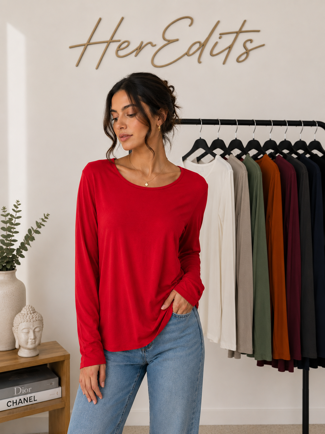 A woman wearing a bright red long-sleeve top and blue jeans stands in a boutique setting with a clothing rack of other colors behind her and a wall sign reading 'HerEdits'.