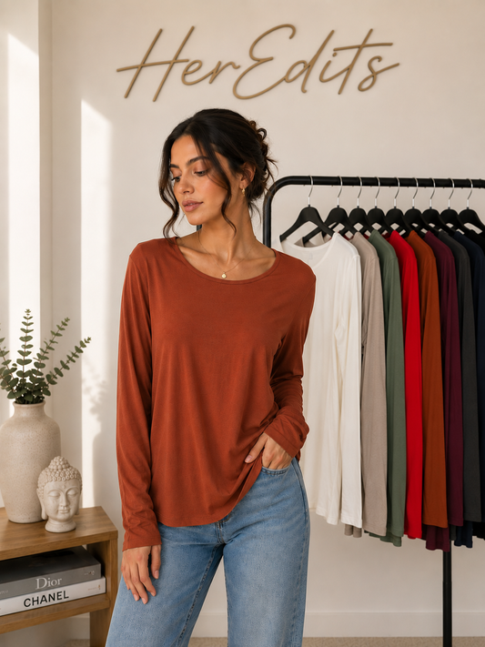 A woman wearing a rust-colored long-sleeve top and light blue jeans stands in a boutique setting with a clothing rack and 'HerEdits' wall sign behind her.