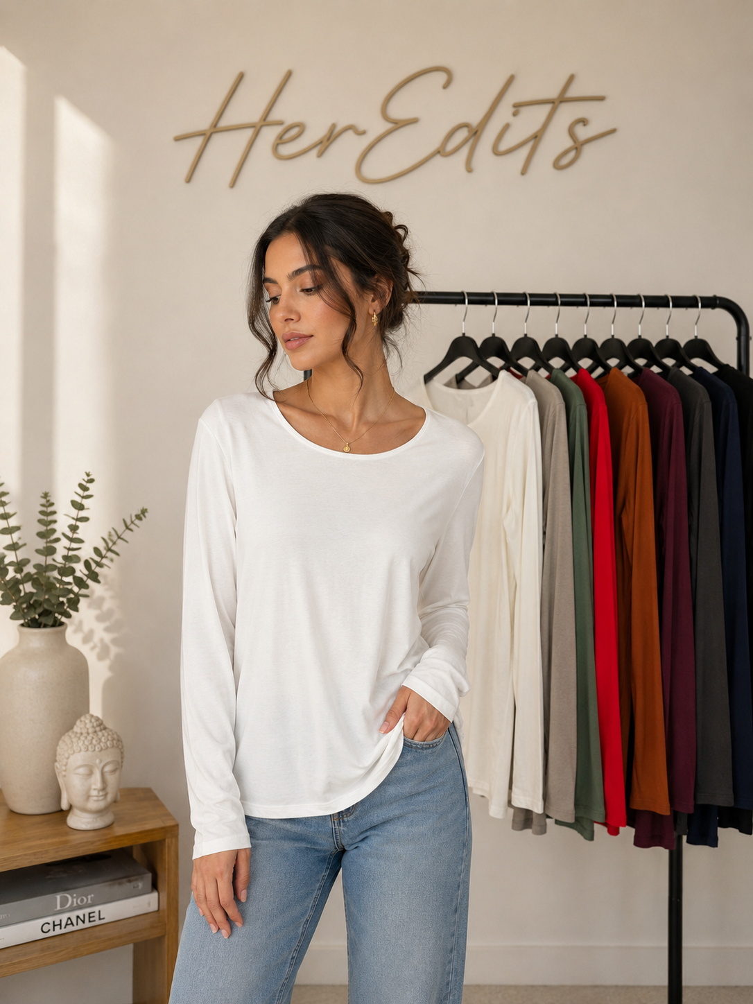 A woman wearing a white long-sleeve crewneck top and light blue jeans stands in a boutique setting. A clothing rack with multiple colored tops is visible behind her, along with a wall sign reading 'HerEdits' and a small wooden side table with decorative items.