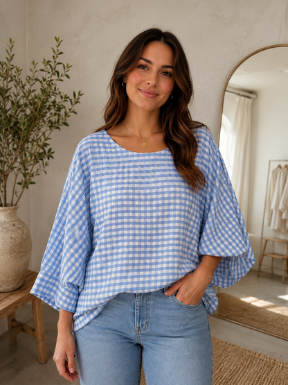 A woman wearing a light blue and white gingham check top with wide batwing sleeves, paired with light-wash blue jeans, standing in a bright, modern room with a potted plant and a mirror in the background.