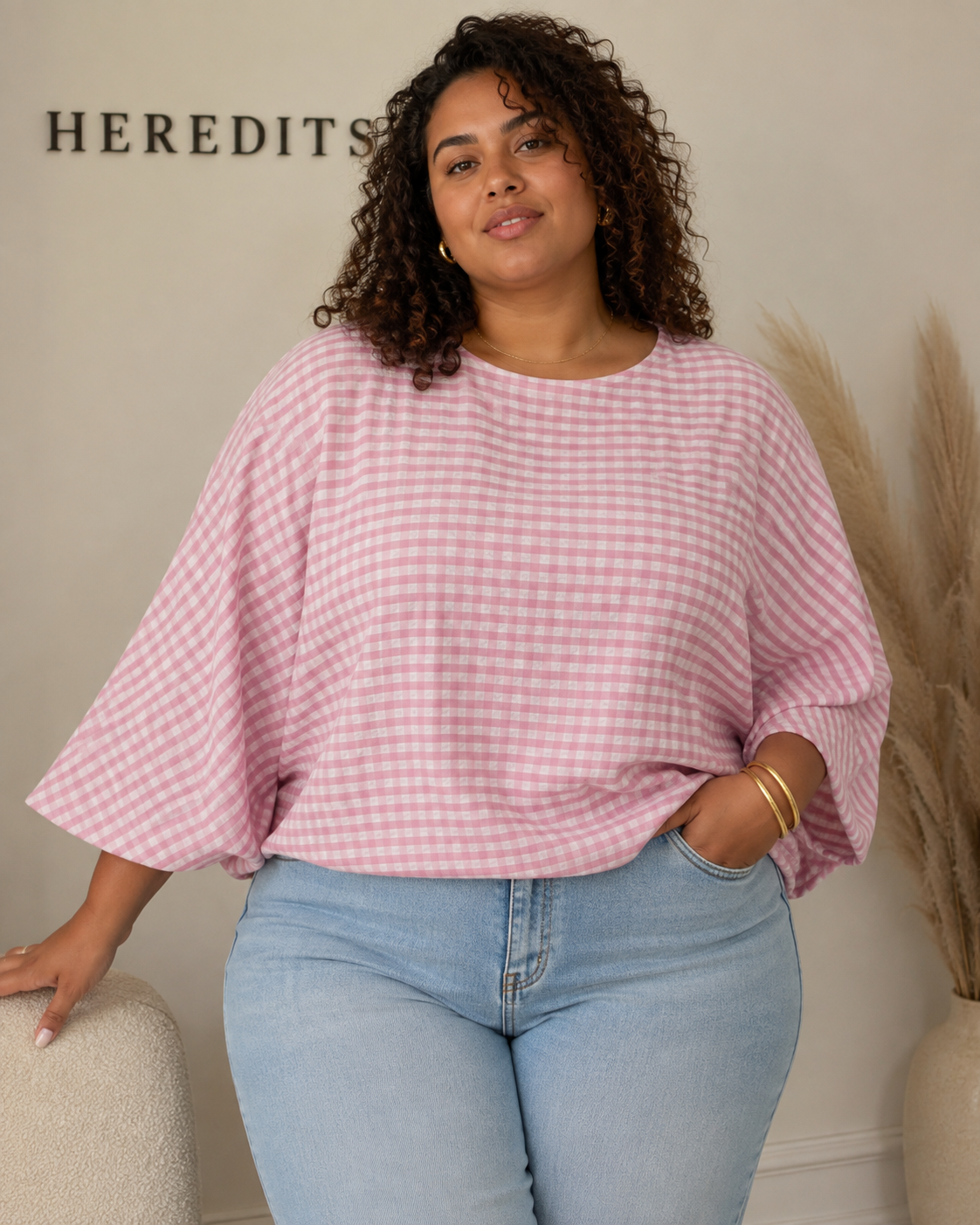 A woman wearing a pink and white gingham check 3/4-sleeve top and light blue jeans, standing indoors against a beige wall with the brand name 'HEREDITIS' partially visible.