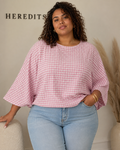 A woman wearing a pink and white gingham check 3/4-sleeve top and light blue jeans, standing indoors against a beige wall with the brand name 'HEREDITIS' partially visible.