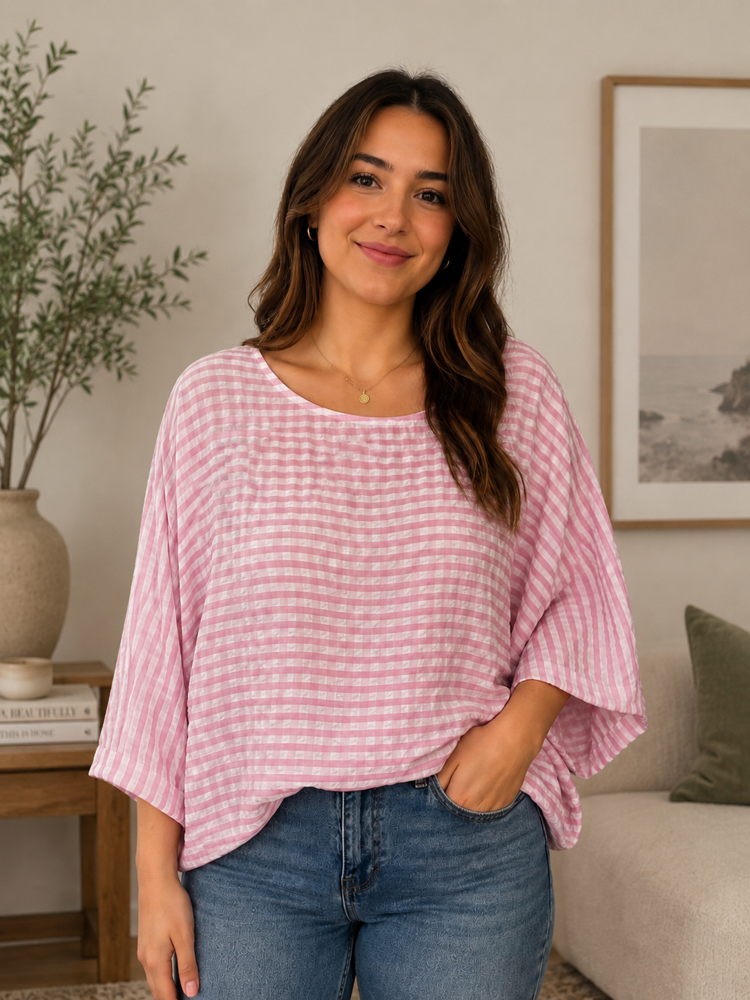 A woman wearing a pink and white gingham patterned batwing top with blue jeans, standing in a cozy living room setting.