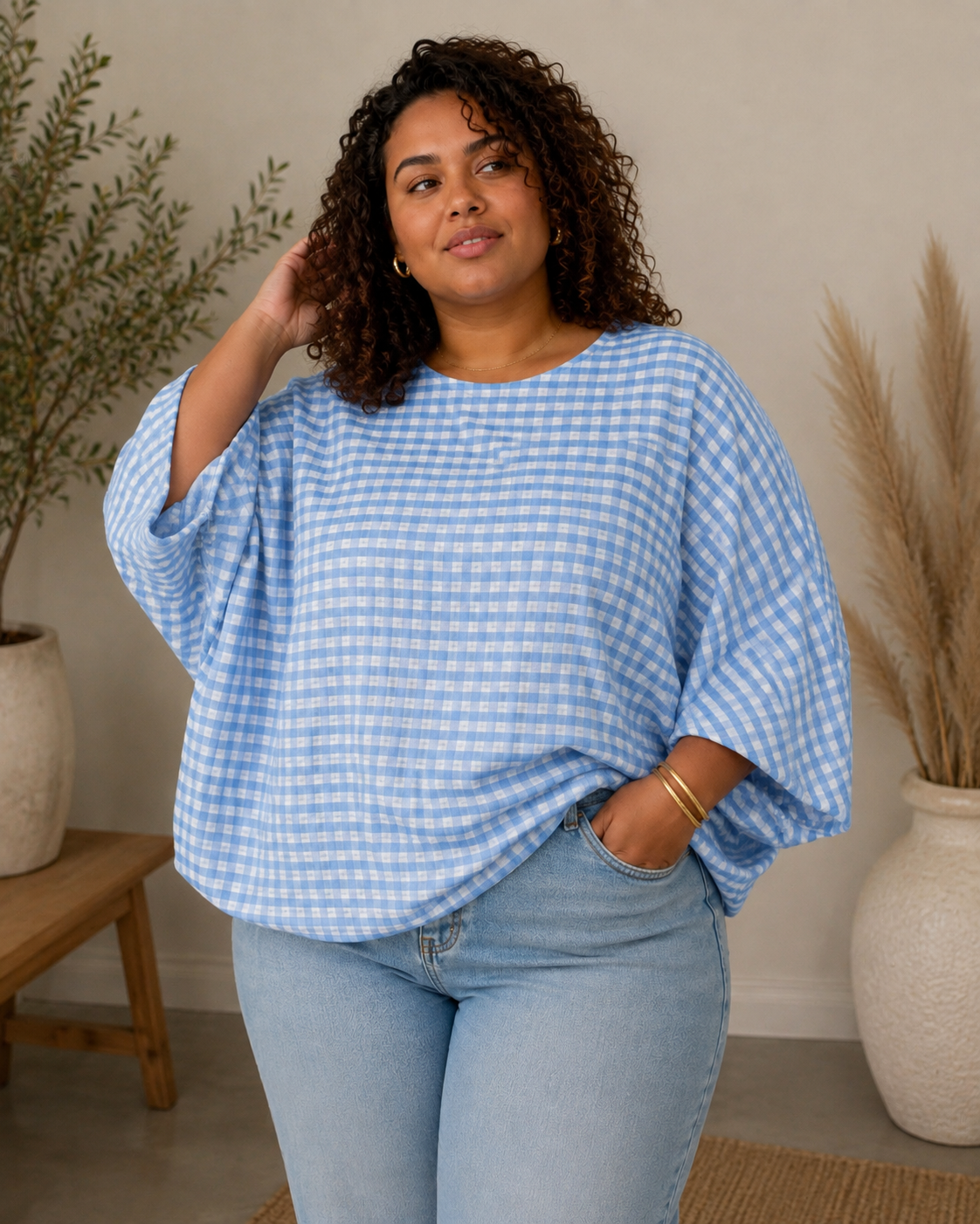 A woman wearing a light blue and white gingham batwing-sleeve top and light-wash jeans, standing in a styled indoor setting with potted plants and a wooden stool.