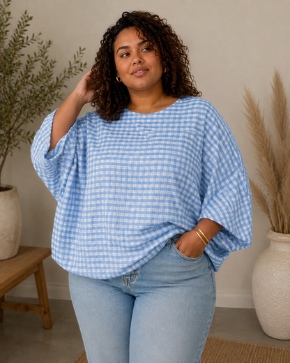 A woman wearing a light blue and white gingham batwing-sleeve top and light-wash jeans, standing in a styled indoor setting with potted plants and a wooden stool.