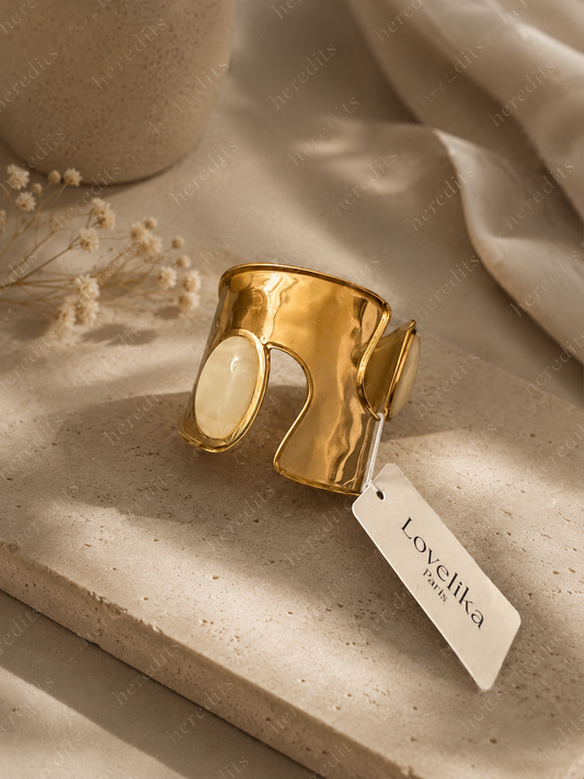 A wide gold-toned cuff ring with a smooth oval cream inlay, styled on a beige stone slab with a fabric tag reading Lovetika Paris.