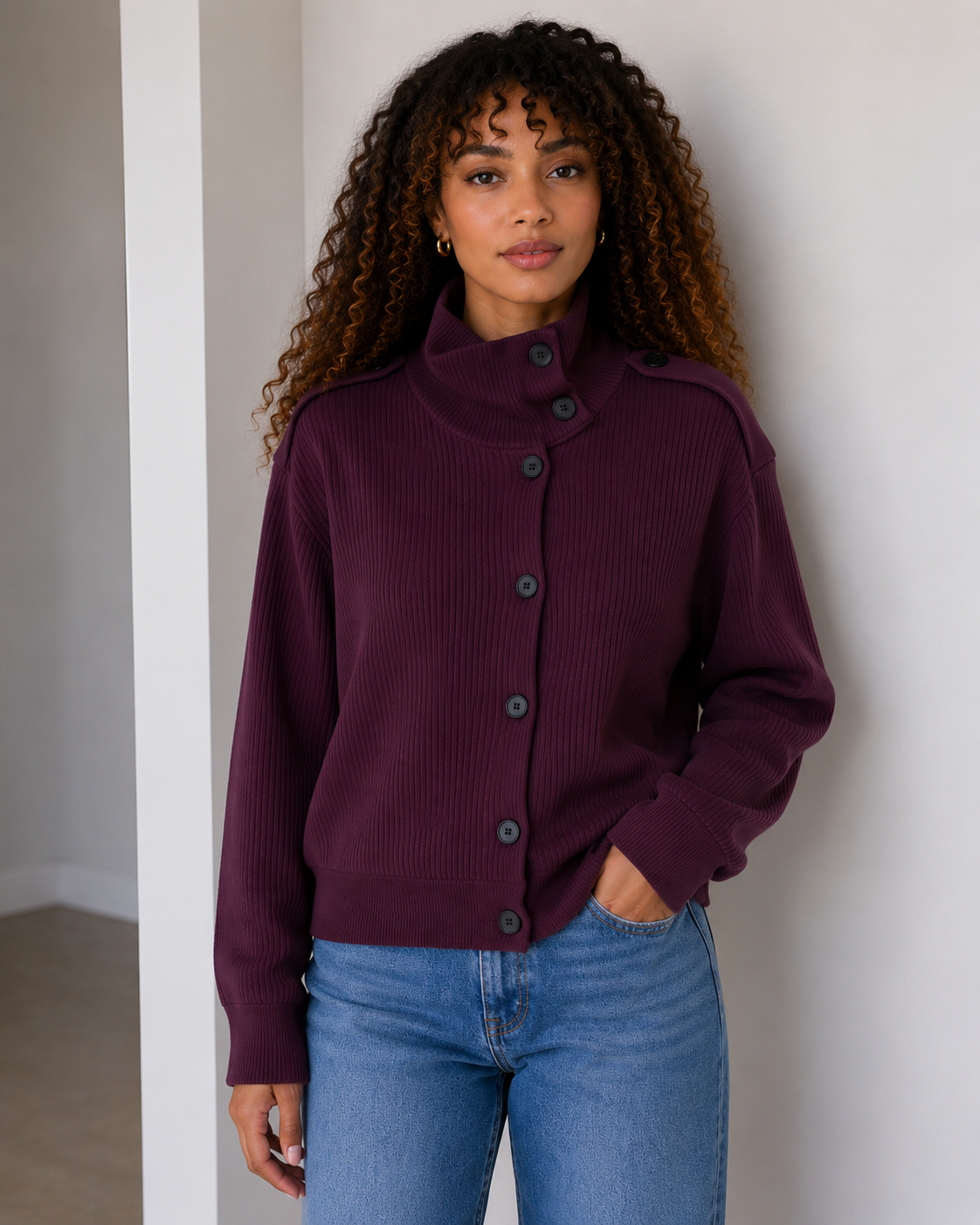 A woman with curly hair wearing a dark purple ribbed button-up cardigan and blue jeans, standing against a white wall.