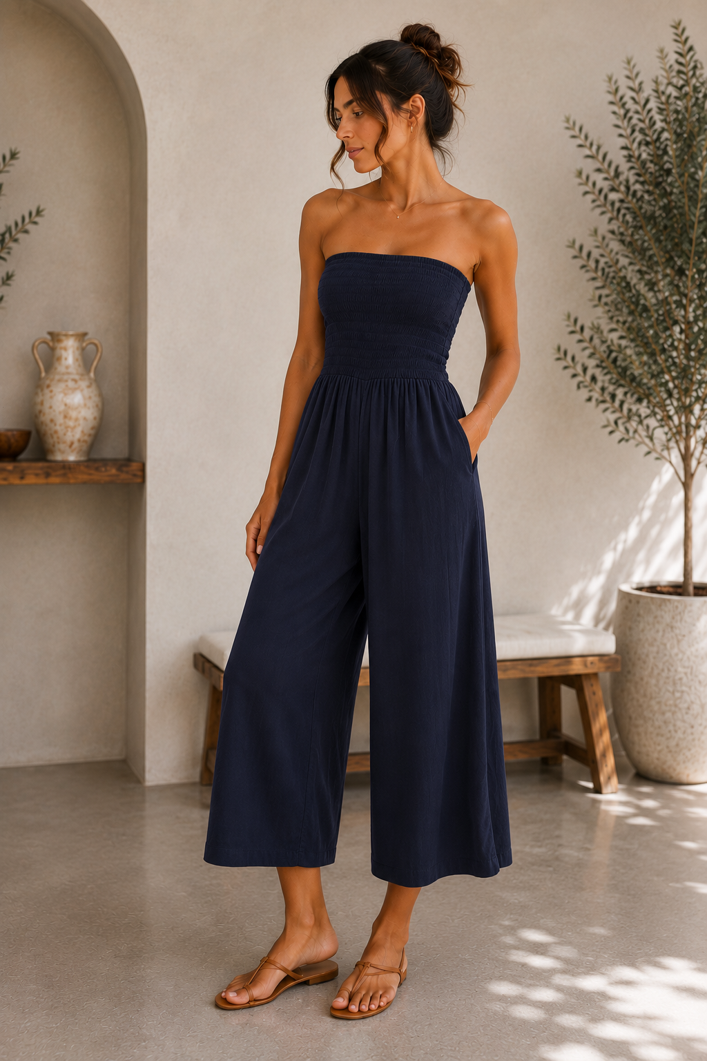 A woman modeling a navy blue strapless wide-leg jumpsuit in a bright, modern interior setting with a concrete floor, arched wall niche, and potted plant.