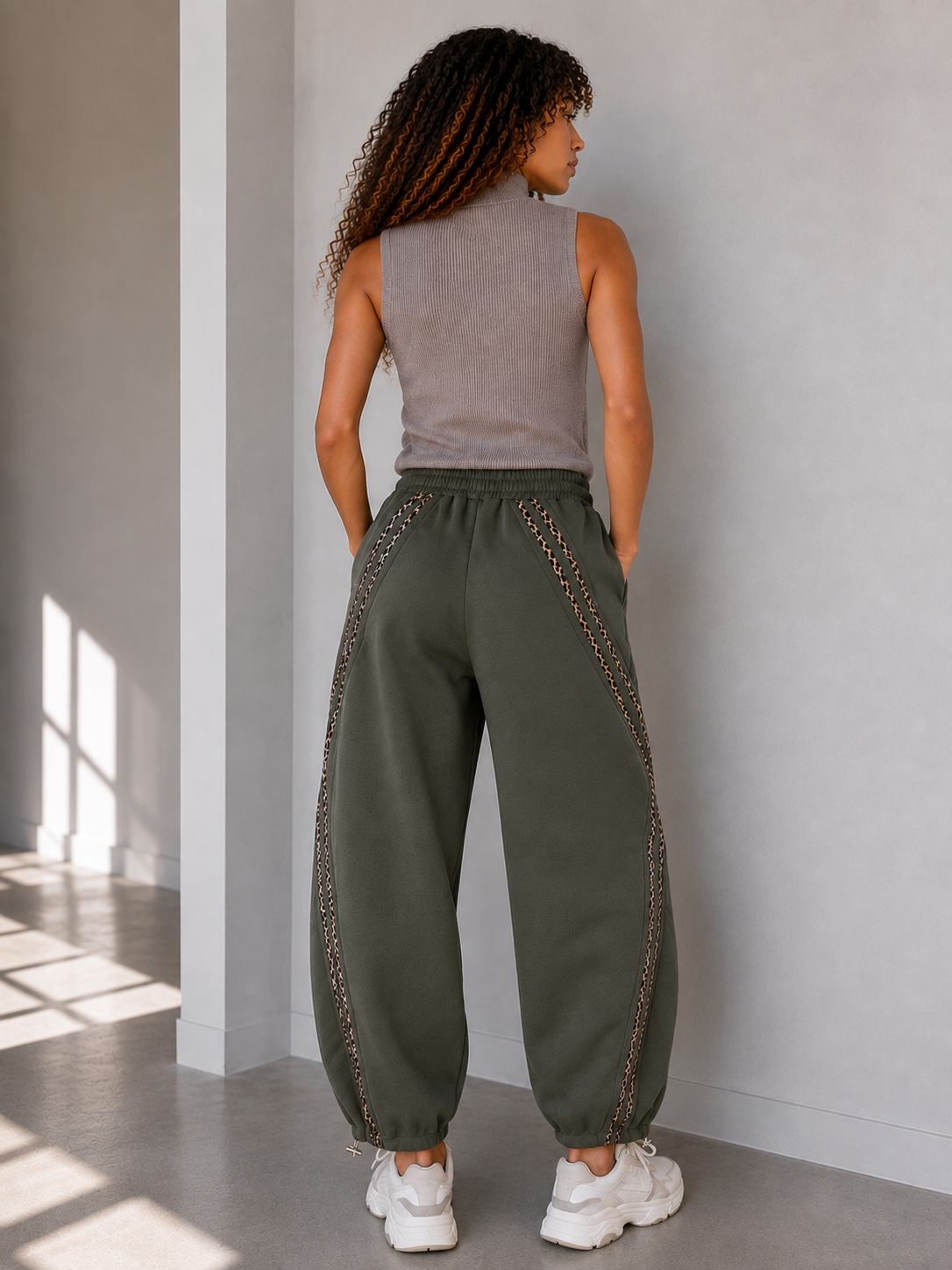 A woman shown from behind wearing loose, wide-leg barrel khaki harem-style pants with decorative chain-like trim along the sides, paired with a sleeveless top and white sneakers in a minimalist indoor setting.
