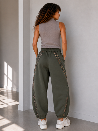 A woman shown from behind wearing loose, wide-leg barrel khaki harem-style pants with decorative chain-like trim along the sides, paired with a sleeveless top and white sneakers in a minimalist indoor setting.