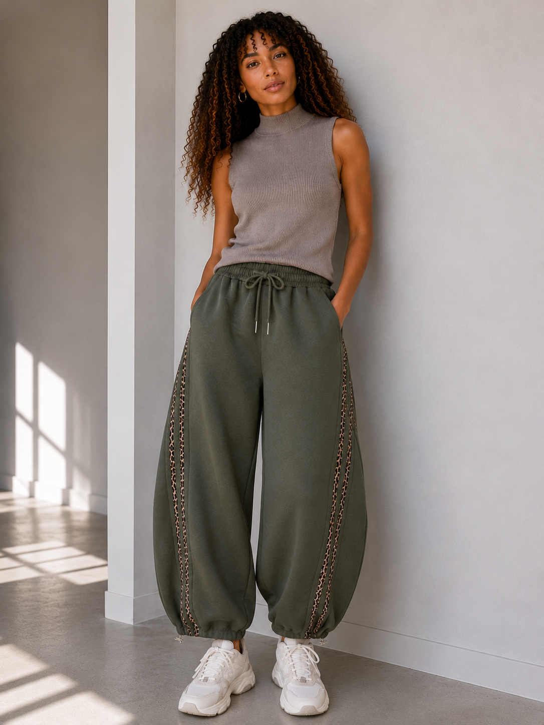 A woman stands indoors against a light grey wall wearing loose  khaki green drawstring pants with decorative chain-like side detailing, a taupe sleeveless top, and white sneakers.