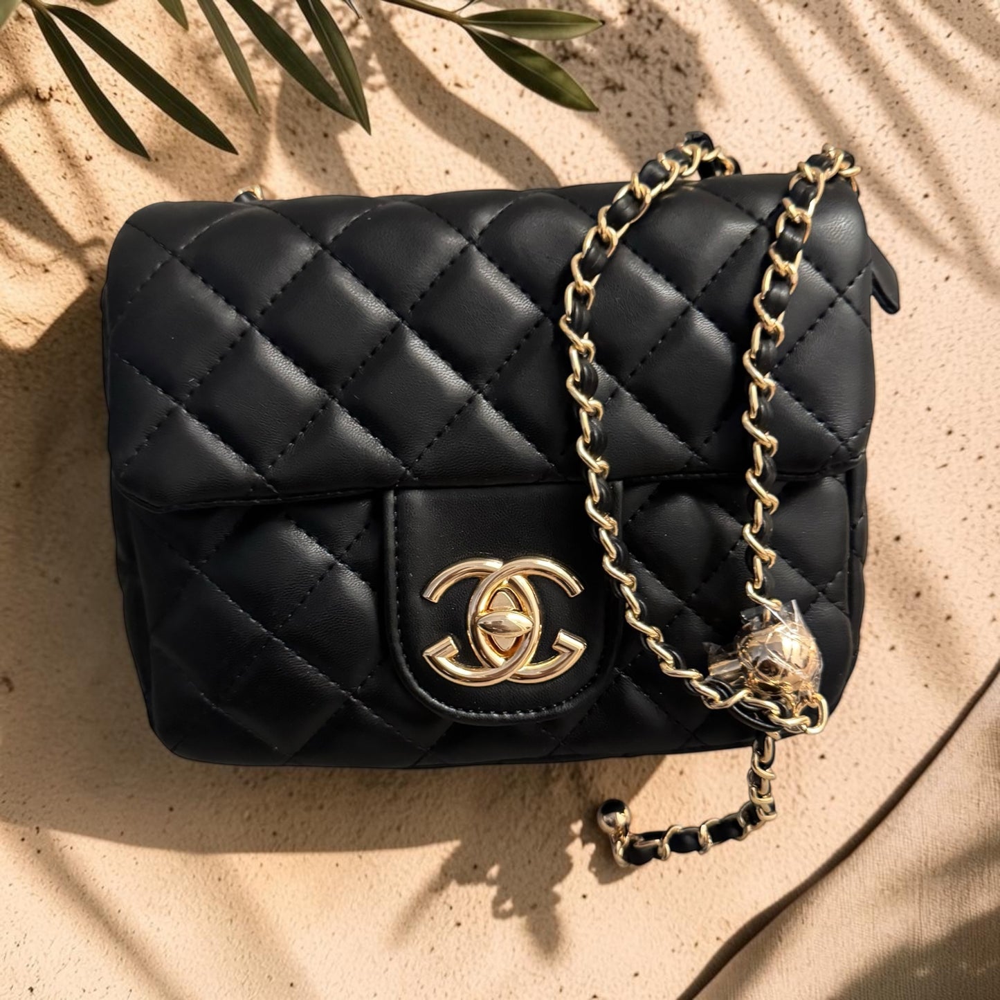 Black quilted handbag with gold chain and emblem on a beige surface with leaf shadows