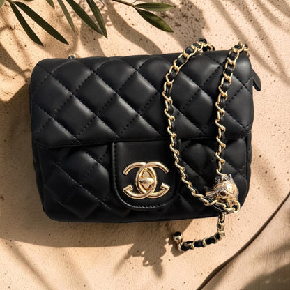 Black quilted handbag with gold chain and emblem on a beige surface with leaf shadows