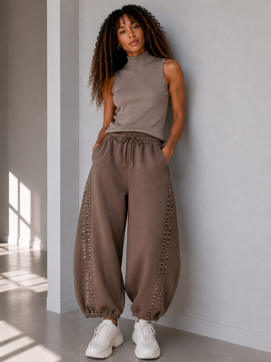 A woman shown from frontwearing loose, wide-leg barrel taupe-brown harem-style pants with decorative chain-like trim along the sides, paired with a sleeveless top and white sneakers in a minimalist indoor setting.