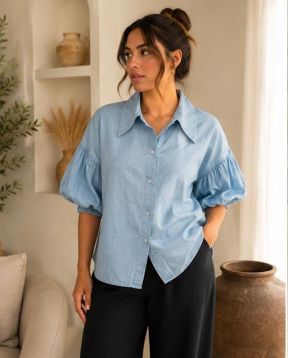 A woman wearing a light blue chambray button-up blouse with puffed sleeves and black pants, standing in a brightly lit room with shelves and a large vase in the background.