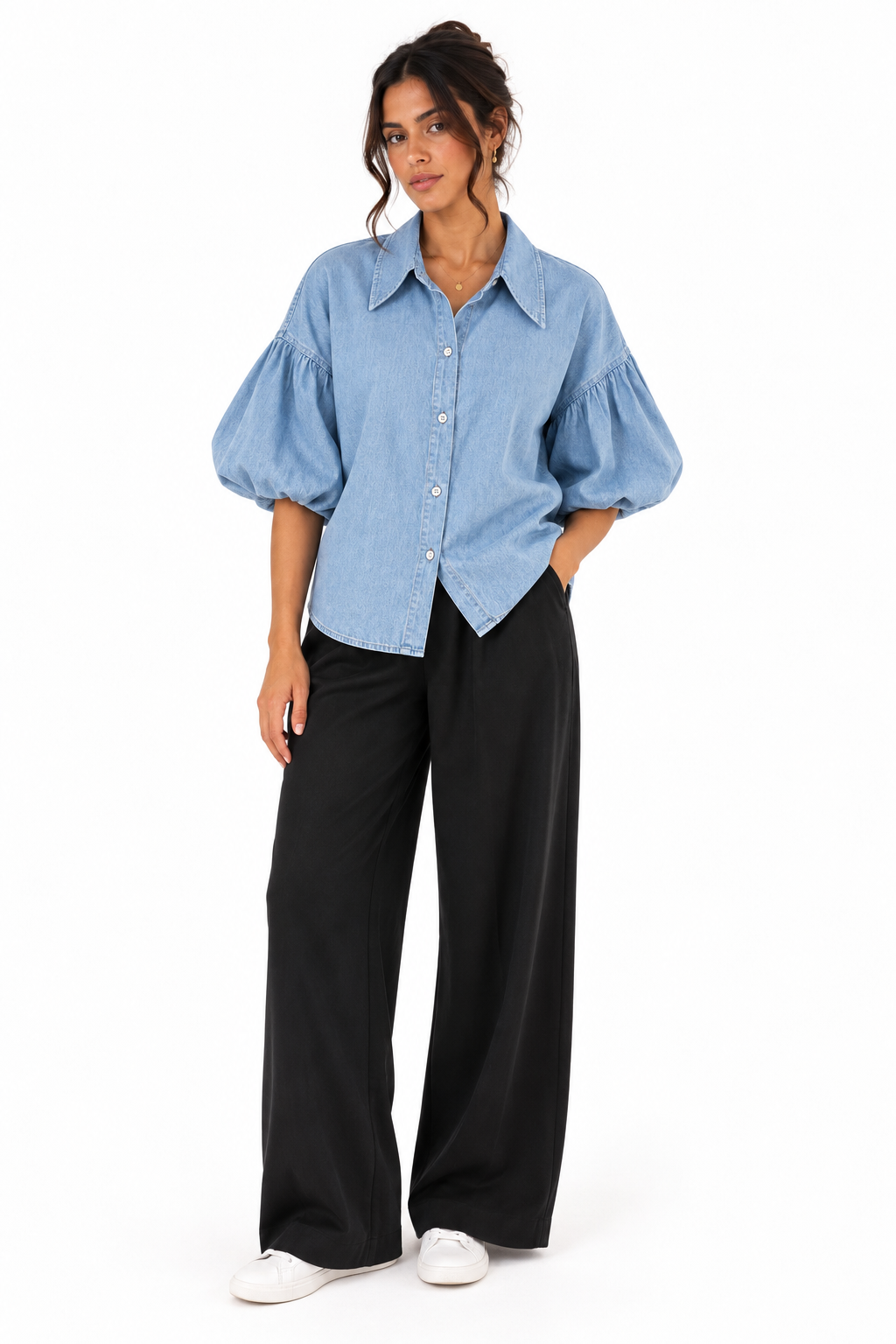 A woman modeling a light blue chambray puff-sleeve button-up shirt paired with black wide-leg trousers and white sneakers, shot against a plain white studio background.