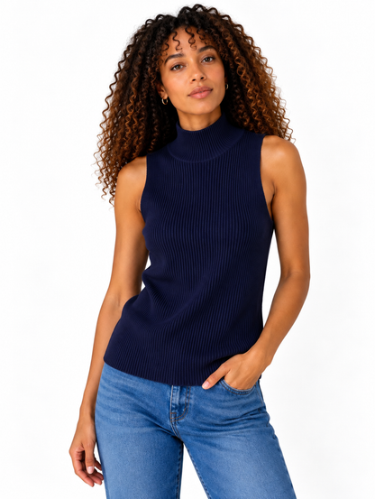 A woman with curly brown hair wearing a navy blue ribbed sleeveless mock neck sweater and blue jeans, standing against a plain white background.