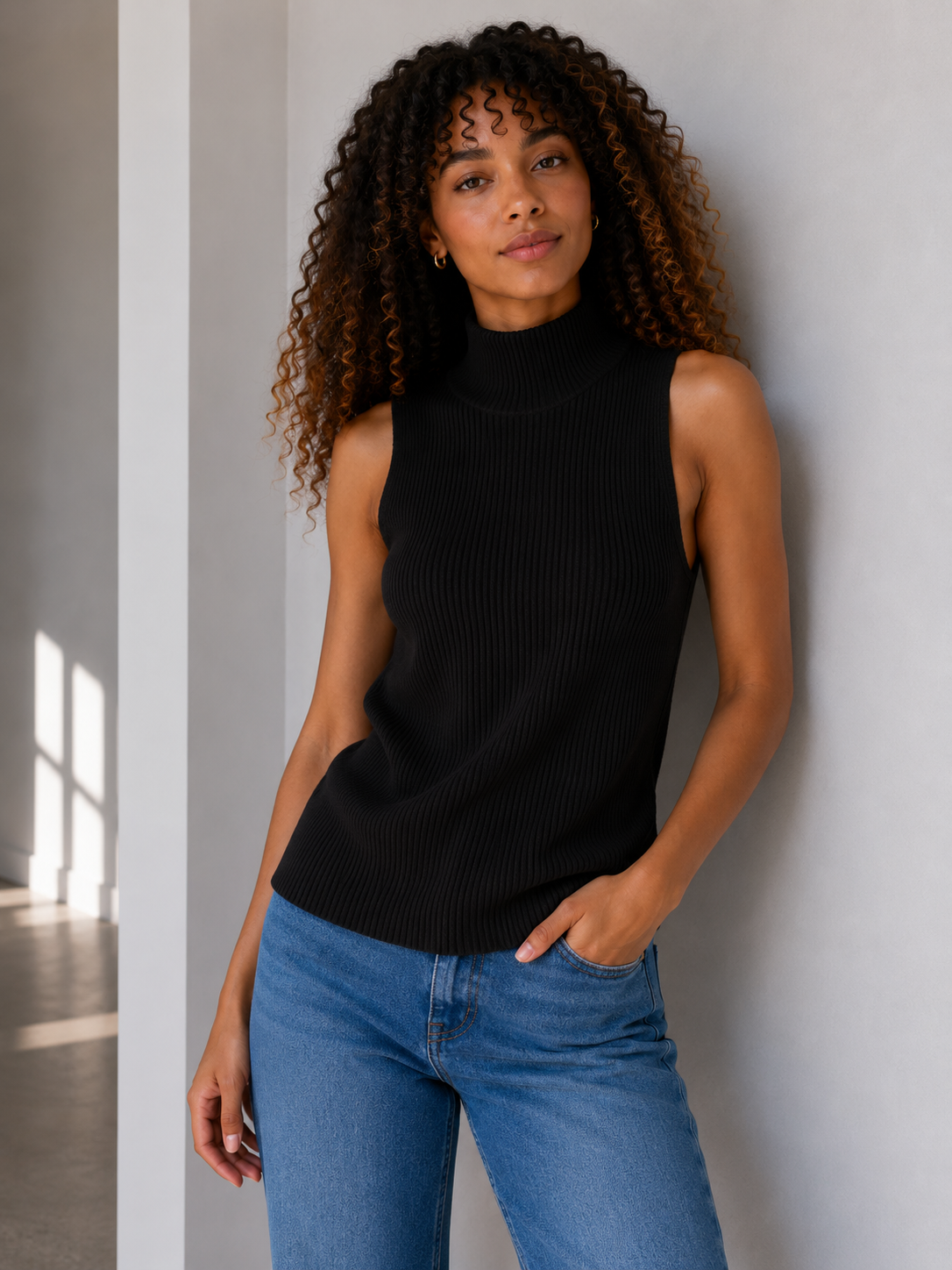 A woman with curly brown hair wearing a sleeveless black ribbed mock neck top and blue jeans, standing against a light gray wall.