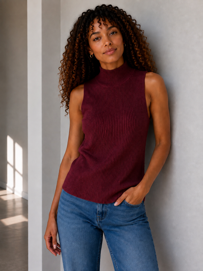 A woman with curly brown hair wearing a sleeveless ribbed mock neck sweater in burgundy and blue jeans, standing against a light gray wall.