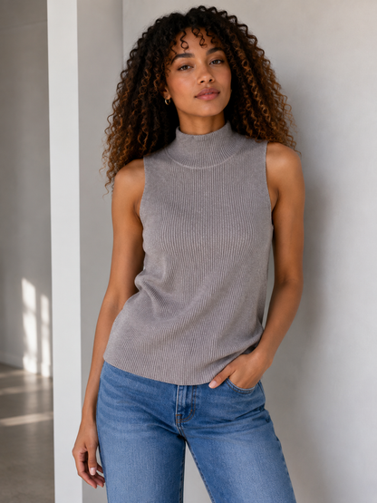 A woman with curly brown hair wearing a sleeveless ribbed grey mock neck top and blue jeans, standing against a light gray wall.