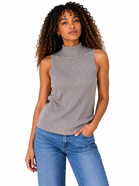 A woman with curly brown hair wearing a sleeveless ribbed grey mock neck top and blue jeans, standing against a light gray wall.
