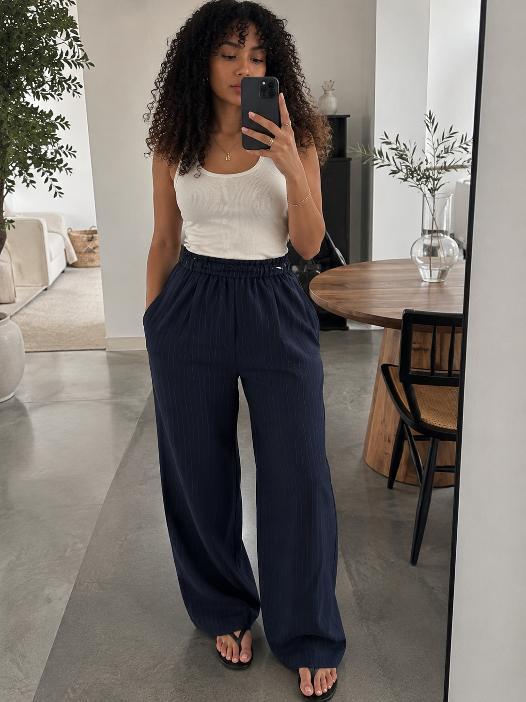 A woman takes a mirror selfie in a modern room, wearing a white ribbed tank top and navy blue wide-leg pinstripe trousers.