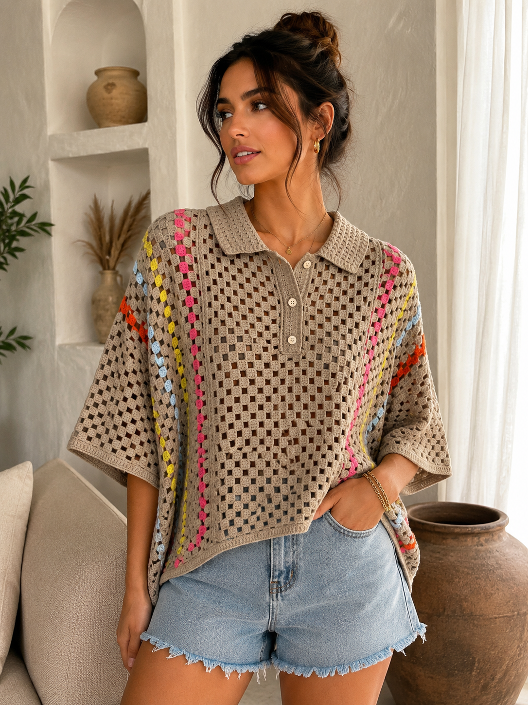 A woman wearing a Mocha crochet knit polo shirt with colorful striped patterns on the sides, paired with light blue denim shorts, standing in a bright, modern room.