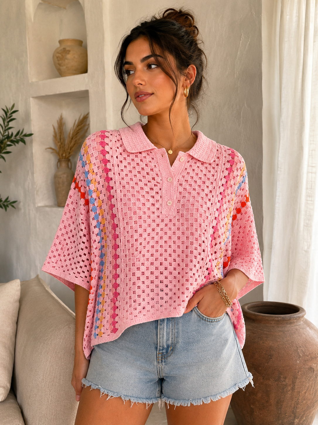 A woman wearing a pink open-knit short-sleeve polo shirt with colorful striped side panels and light-wash denim shorts, standing in a bright indoor room with neutral decor.