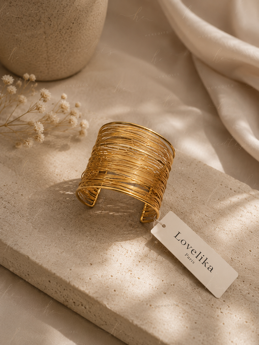 A wide gold wire-wrapped cuff bracelet displayed on a textured beige stone slab with a small white brand tag reading 'Loveliika Paris'. The scene is styled with dried flowers, a ceramic vase, and draped cream fabric under warm, dappled sunlight.