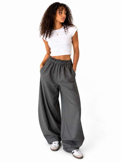 A woman modelling a pair of loose, wide-leg grey pants with an elastic waistband, paired with a white crop top and white sneakers, against a plain white background.