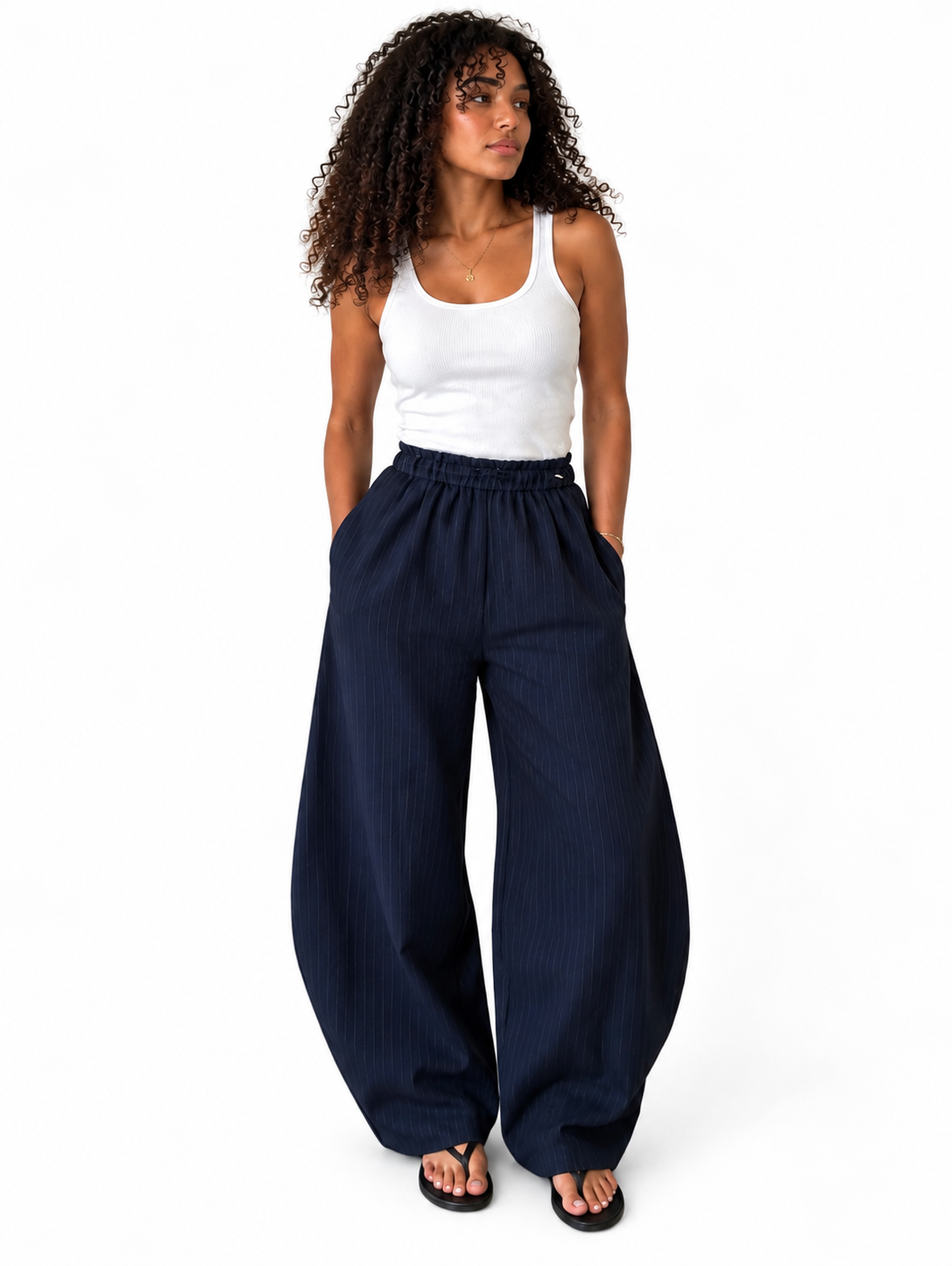 A woman modeling a pair of loose-fitting, wide-leg navy blue pants with a white ribbed tank top and black sandals, standing against a plain white studio background.