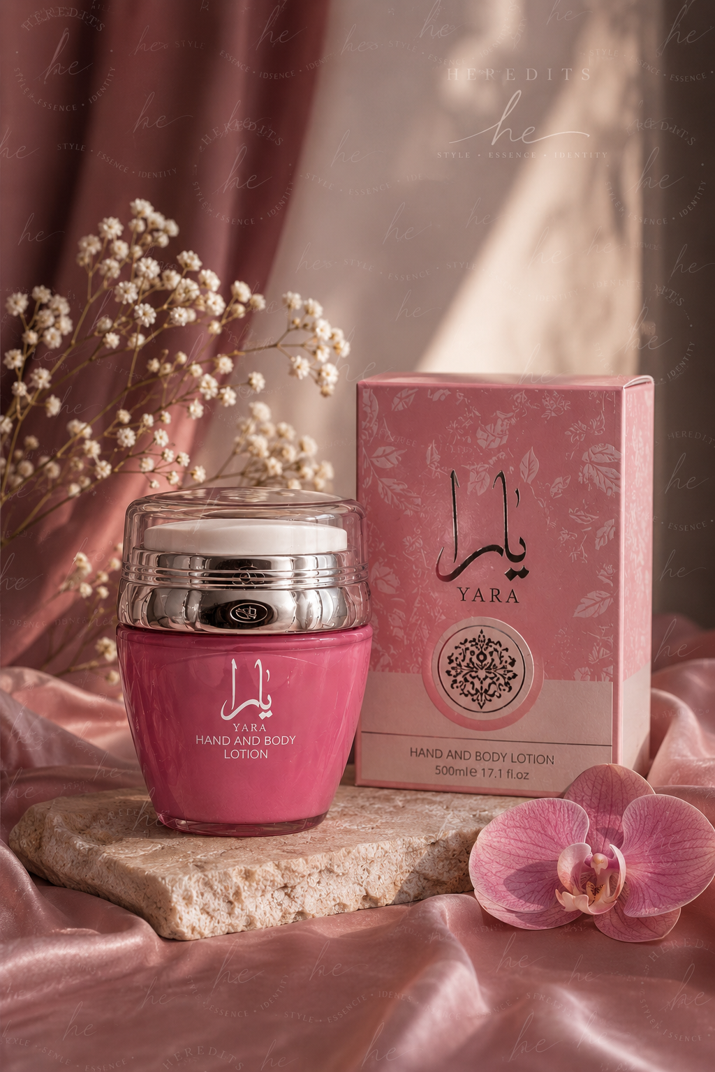 A pink jar of YARA Hand and Body Lotion with a silver lid sits next to its matching pink box on a stone slab, styled with pink satin fabric, baby's breath flowers, and a pink orchid.