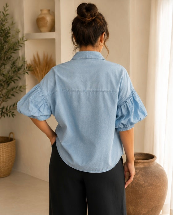 A woman viewed from the back wearing a light blue chambray puff-sleeve blouse and black wide-leg pants in a warm, neutral-toned room.